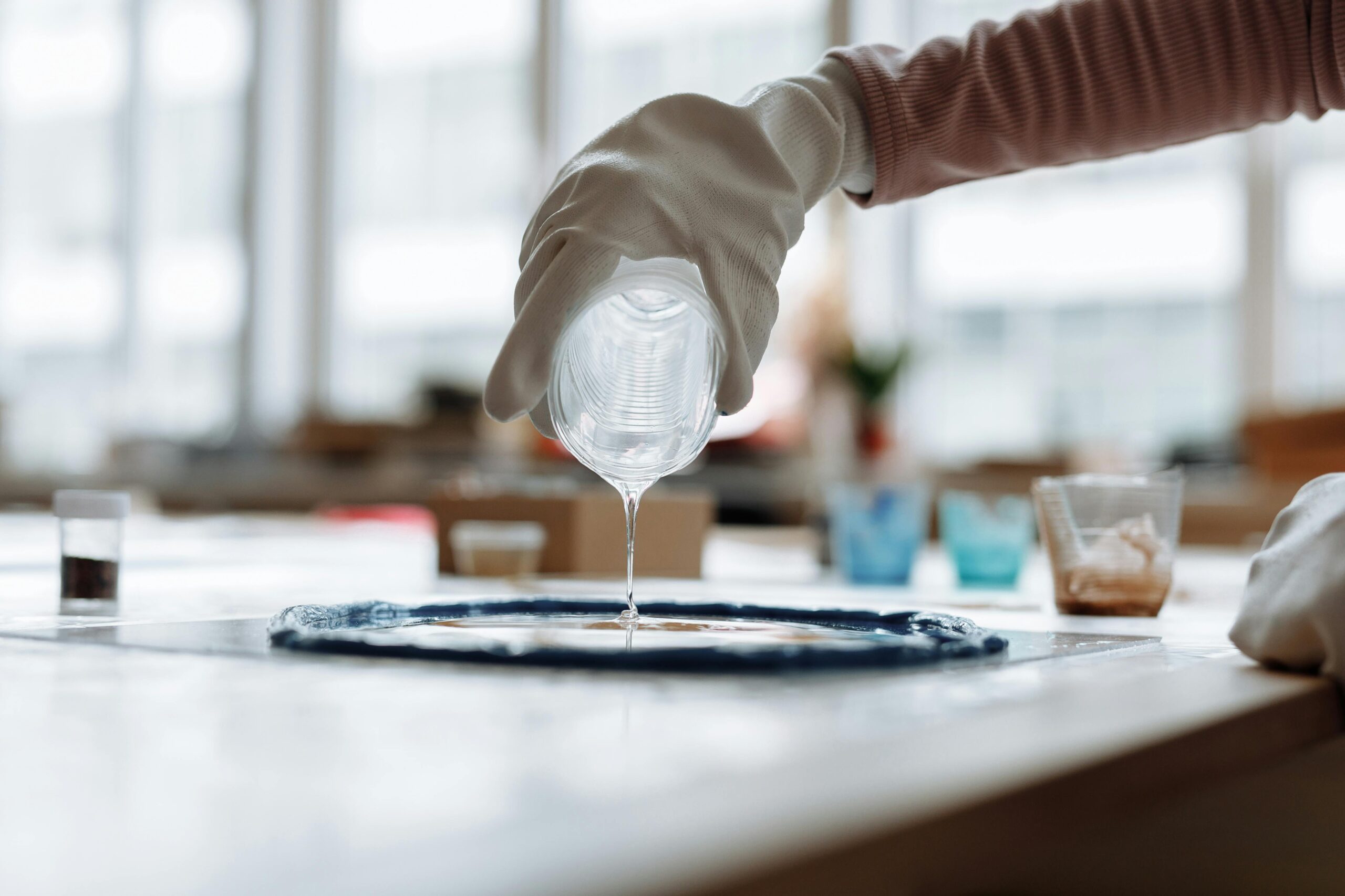 Close-up of a gloved hand pouring clear resin on a table in an indoor art studio.